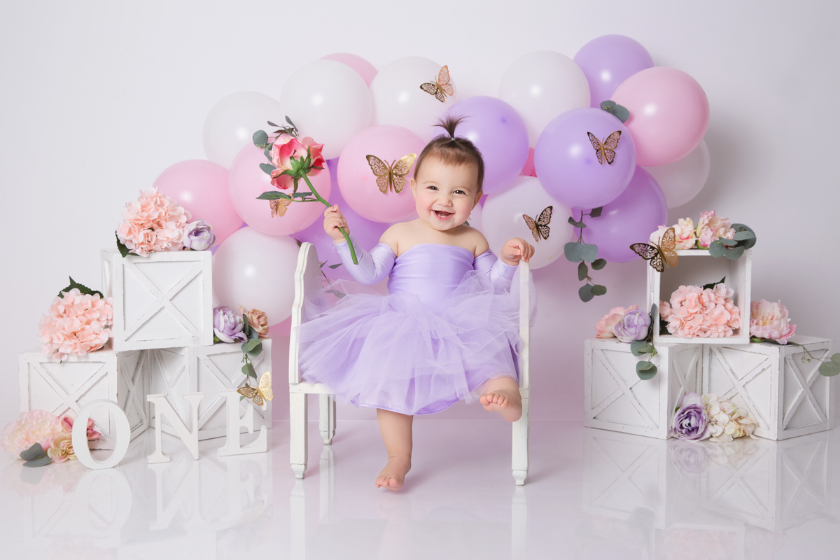 12 month old baby girl in a purple dress sitting on bench holding a flower and smiling surrounded by pink and purple balloons