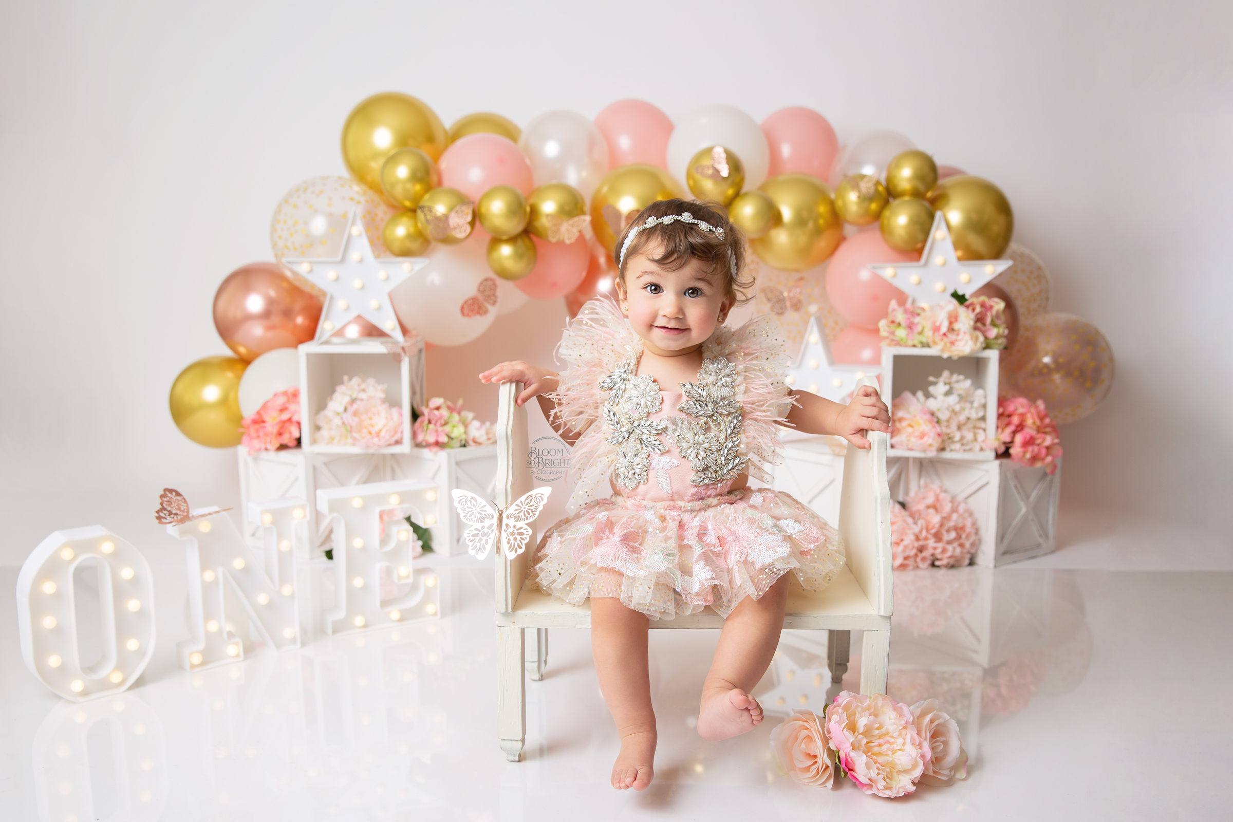 One-year old girl sitting in front of gold, pink and white themed balloons.